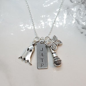 Puede incluir: Collar de plata con varios colgantes, incluyendo un fantasma, un micrófono, una mariposa y una etiqueta rectangular con las letras "JATP" grabadas en ella.