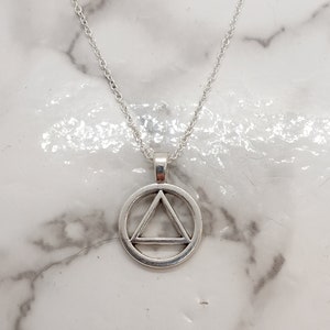 Puede incluir: Un collar de plata con un colgante circular que presenta un triángulo en su interior. El triángulo tiene una línea en el medio.