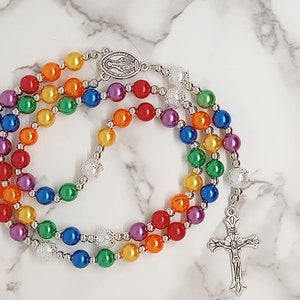 May include: A rainbow rosary with silver beads and a silver crucifix. The beads are made of iridescent glass and are arranged in the colours of the rainbow.