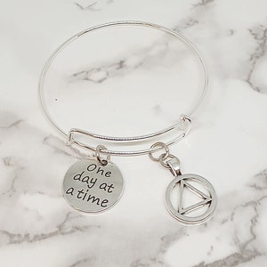 Può includere: Braccialetto in argento con due ciondoli. Un ciondolo è un cerchio con un triangolo all'interno e l'altro ciondolo è un disco rotondo con il testo "One day at a time".