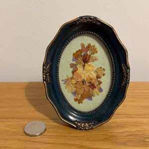 Puede incluir: Un marco ovalado de color verde azulado oscuro con detalles dorados rodea un arreglo floral prensado sobre un fondo verde claro. El marco está sobre una superficie de madera, con una moneda visible cerca.