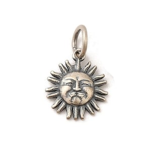 May include: A silver sun charm with a smiling face and radiating rays. The charm has a small loop at the top for attaching to a necklace or bracelet. The sun's face has detailed features, including a moustache.