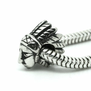 Puede incluir: Un abalorio de plata con un diseño detallado de un jefe nativo americano con un tocado de plumas. El abalorio está diseñado para encajar en una pulsera de abalorios.