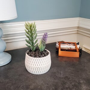 May include: A white ceramic planter with a geometric pattern, filled with black pebbles and a small succulent plant with green leaves and purple flowers.