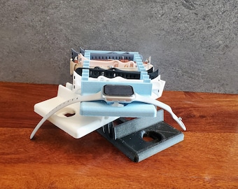 May include: A 3D printed watch stand with a light blue, white, and black design. The stand is holding a silver smartwatch with a black band.