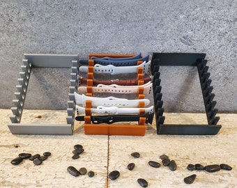 May include: Three watch band display stands in gray, orange, and black, each with a series of angled slots to hold watch bands. The stands hold various colored watch bands, including blue, white, and brown leather. Small, dark stones are scattered in front.