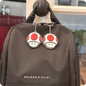 May include: Two mushroom-shaped keychains with red spots and white caps hang from a black backpack. The backpack has the brand name "BRIGGS & RILEY" printed on it. The keychains are attached to the zipper of the backpack.
