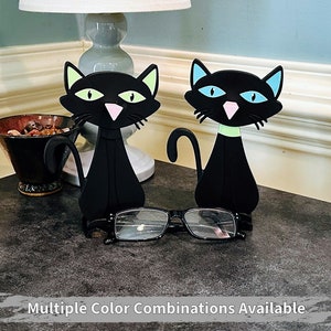 May include: Two black cat-shaped eyeglass holders with green and blue accents. The cats are standing on a dark gray surface with a pair of eyeglasses in front of them. Multiple Color Combinations Available.
