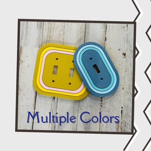 May include: Two light switch covers, one square and one oval, with multiple colored stripes. The square cover is yellow with pink and black stripes. The oval cover is blue with light blue and white stripes. The text "Multiple Colors" is below the covers.