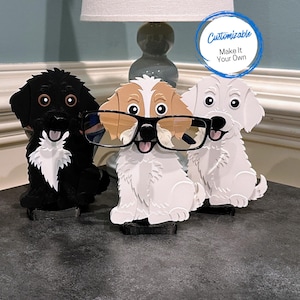 May include: Three dog-shaped eyeglass holders in black, white, and tan. The center dog wears glasses. The holders are on a dark surface. A blue circle with the text "Customizable Make It Your Own" is in the background.