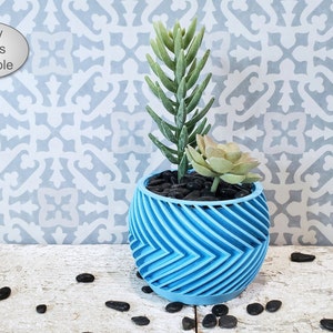 May include: A blue planter with a geometric pattern, holding a small succulent plant and a larger green plant. The planter is sitting on a light wood surface with black stones scattered around it. "Many Colors Available" is written on a white oval sticker in the top left corner of the image.