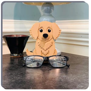 May include: A wooden golden retriever dog figurine holding a pair of black spectacles. The figurine is sitting on a dark surface.