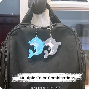 May include: Two dolphin keychains, one light blue and one gray, hang from a black bag. The keychains are attached to silver chains. The text "Multiple Color Combinations" is visible at the bottom of the image.