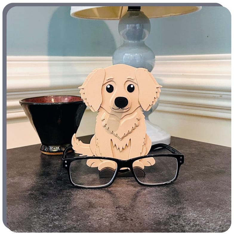 May include: A wooden figurine of a golden retriever puppy sitting on a table with a pair of spectacles resting on its back. The puppy has a light brown coat and black eyes. The spectacles are black with clear lenses.