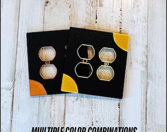 May include: Two black electrical outlet covers with gold and orange corner accents. The covers have octagonal cutouts and gold-colored outlines. The text "MULTIPLE COLOR COMBINATIONS" is at the bottom of the image.