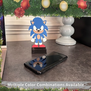 May include: A blue and red Sonic the Hedgehog figurine standing on a black base. The figurine is on a gray surface with a black smartphone. The image is framed with a green garland with red and gold ornaments.