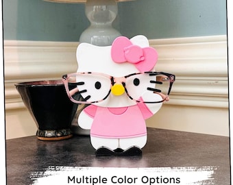 Puede incluir: Un personaje de Hello Kitty rosa con gafas negras y un lazo rosa. El personaje es un soporte para gafas.