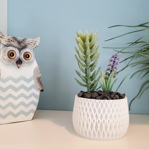 May include: A white ceramic planter with a geometric pattern holds a small artificial succulent plant with purple flowers. A wooden owl figurine with a blue and white chevron pattern is on the left side of the image.