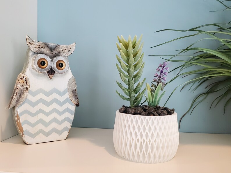 May include: A white ceramic planter with a geometric pattern holds a small artificial succulent plant with purple flowers. A wooden owl figurine with a blue and white chevron pattern is on the left side of the image.