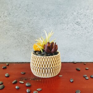 May include: A small, decorative planter made of woven wood with a geometric pattern. The planter is filled with black pebbles and artificial succulents in shades of yellow, green, and burgundy.