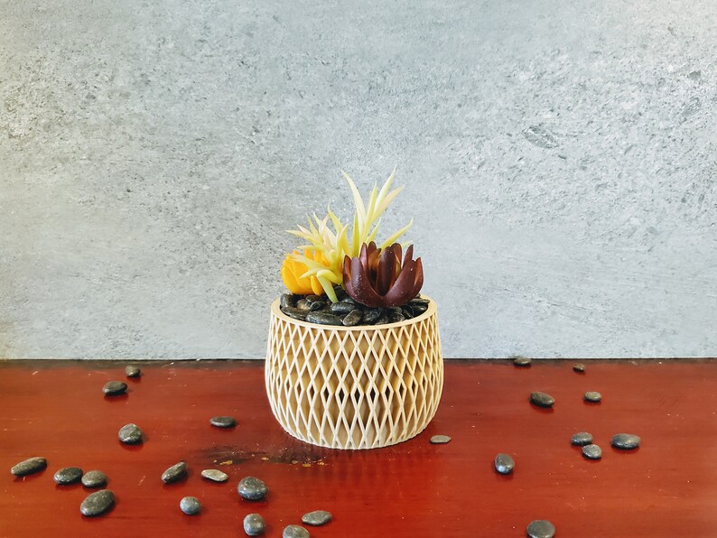 May include: A small, decorative planter made of woven wood with a geometric pattern. The planter is filled with black pebbles and artificial succulents in shades of yellow, green, and burgundy.