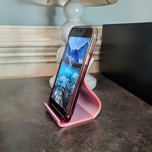 May include: A pink phone stand with a smartphone in a clear case resting on top. The phone screen shows a landscape image of a lake and mountains.