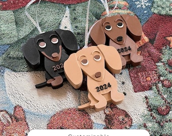 Puede incluir: Tres adornos de dachshund, uno negro, uno marrón y uno marrón claro, con el año 2024 impreso en cada uno. Los adornos están colgados de un árbol de Navidad.