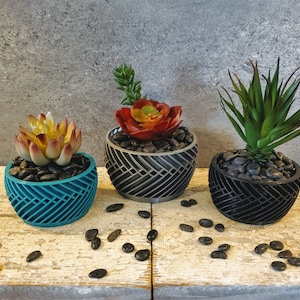 May include: Three geometric patterned planters with black stones and artificial succulents. The planters are teal, gray, and black.