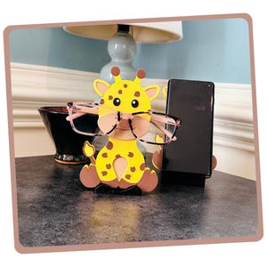 May include: A yellow and brown giraffe-shaped phone and glasses holder. The giraffe is wearing pink glasses and has brown spots. The holder is sitting on a dark gray surface.