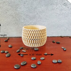 May include: A light brown wooden planter with a diamond pattern design. The planter is 4.35 inches wide and 2.95 inches high. The planter is sitting on a red wooden surface with black stones scattered around it.