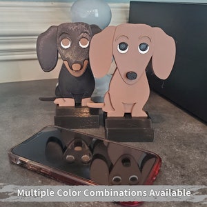 May include: Two wooden dachshund figurines, one brown and one black, with painted details. The figurines are standing on black bases. The figurines are on a gray surface with a cell phone in the foreground.