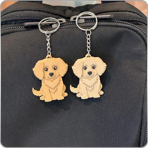 May include: Two gold-coloured dog keyrings with silver key rings hanging from a black backpack. The dogs are sitting with their paws together and have brown eyes.