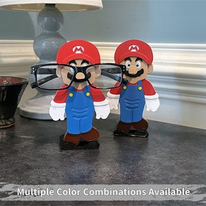 May include: Two red and blue Mario character figurines holding black glasses. The figurines are standing on a dark gray surface.