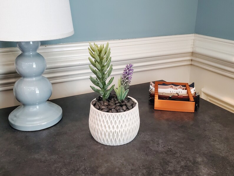 May include: A white ceramic planter with a geometric pattern, filled with black pebbles and a small succulent plant with green leaves and purple flowers.