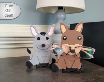 May include: Two cute donkey-shaped eyeglasses holders. One is gray and the other is brown. Both have black eyes and a black nose. The brown donkey is holding a pair of glasses.