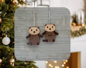 May include: Two brown wooden ornaments shaped like otters hanging from a gray background. The otter on the right has the year 2023 written on it.