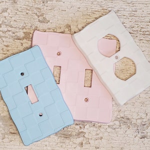May include: Three ceramic light switch and outlet covers. One is white with a double outlet, one is pink with a single switch, and one is light blue with a single switch. All have a textured brick pattern.