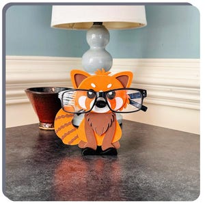 May include: A red panda figurine with black glasses, holding a pair of glasses. The figurine is made of felt and has a brown nose and black eyes. The figurine is sitting on a dark brown surface.