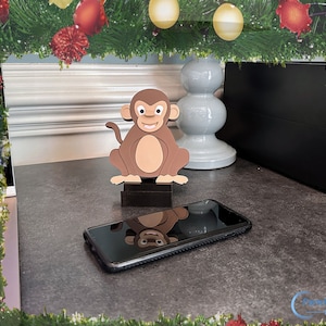 May include: A brown monkey figurine with a black base, sitting on a dark gray surface with a black smartphone in front of it. The monkey has a smiling face and is holding its hands together. The background is a blurry image of a Christmas garland with red and gold ornaments.