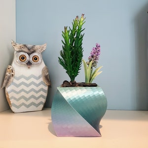 May include: A teal and purple geometric planter with a textured surface holds a small green succulent and a purple flower.