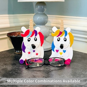 May include: Two white unicorn-shaped eyeglass holders with colorful details. One unicorn has pink, purple, and blue accents with stars, while the other has yellow, pink, and blue accents with hearts. The unicorns are holding a pair of eyeglasses.