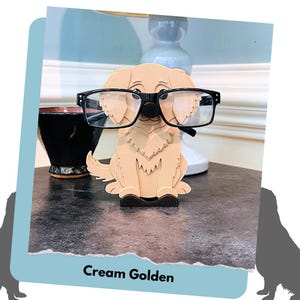 May include: A cream-coloured wooden figurine of a golden retriever wearing black glasses. The figurine is a glasses holder and is sitting on a dark grey surface.