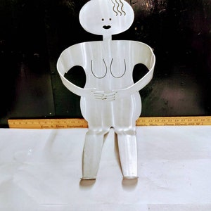 Può includere: Una scultura in metallo argentato di una persona con un design semplice e stilizzato. La scultura è in piedi con le braccia incrociate e ha una linea ondulata per i capelli.