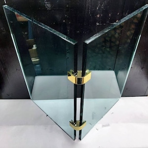 May include: A glass fireplace screen with two panels and gold-colored metal hinges. The screen is made of clear glass and has a triangular shape.