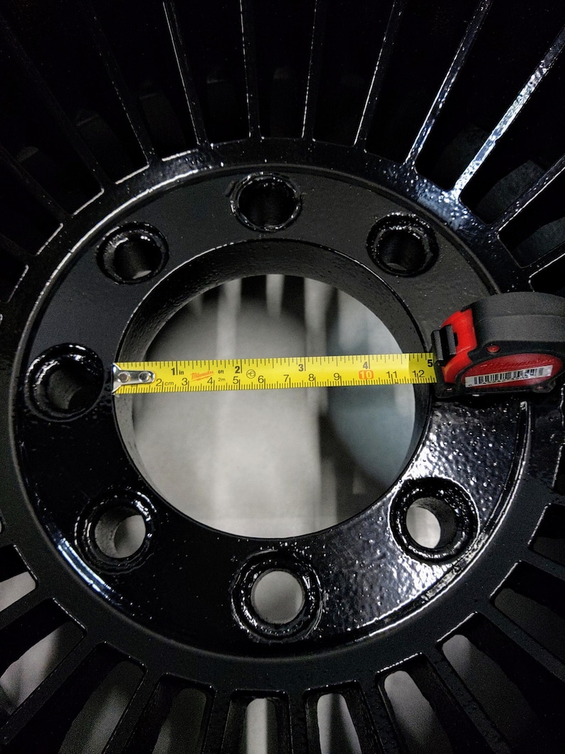 Puede incluir: Una rueda de metal negro con una abertura circular en el centro. La abertura se mide con una cinta m&eacute;trica amarilla, mostrando un di&aacute;metro de 12,7 cm. La rueda tiene una serie de radios que irradian desde el centro.