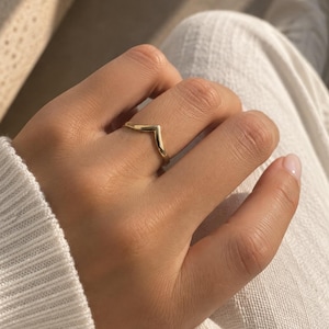 May include: A polished gold ring featuring a V-shaped chevron design. The band has a smooth, reflective surface and a pointed center peak, creating a simple geometric silhouette that sits comfortably on the finger with a bright metallic finish.