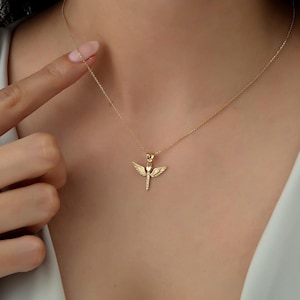 May include: A gold necklace with a small angel wing pendant. The pendant has a heart in the center and is adorned with small sparkling stones.