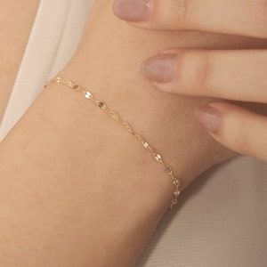 May include: A delicate gold chain bracelet with small, flat, oval-shaped links. The bracelet is shown on a person's wrist.