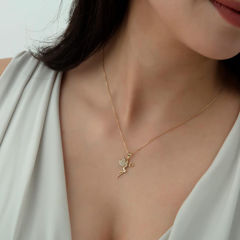 May include: A gold chain necklace with a small pendant featuring a stylized figure with wings and a heart. The pendant is adorned with small, clear stones.