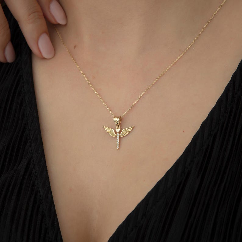 May include: A gold necklace with a small angel pendant featuring a heart and wings. The angel is adorned with small clear stones.